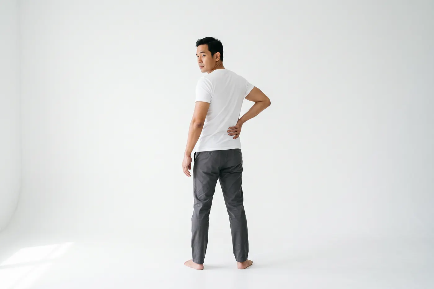 Back view of a man in a white studio turning to look over one shoulder, one hand pressed to his lower back