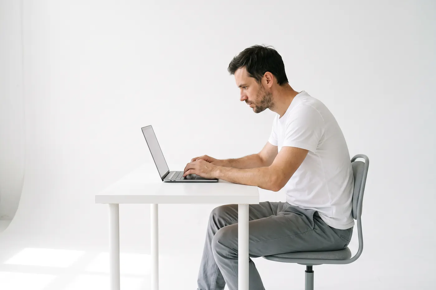 Side view of a man seated at a laptop in a white studio, his head and neck angled forward toward the screen