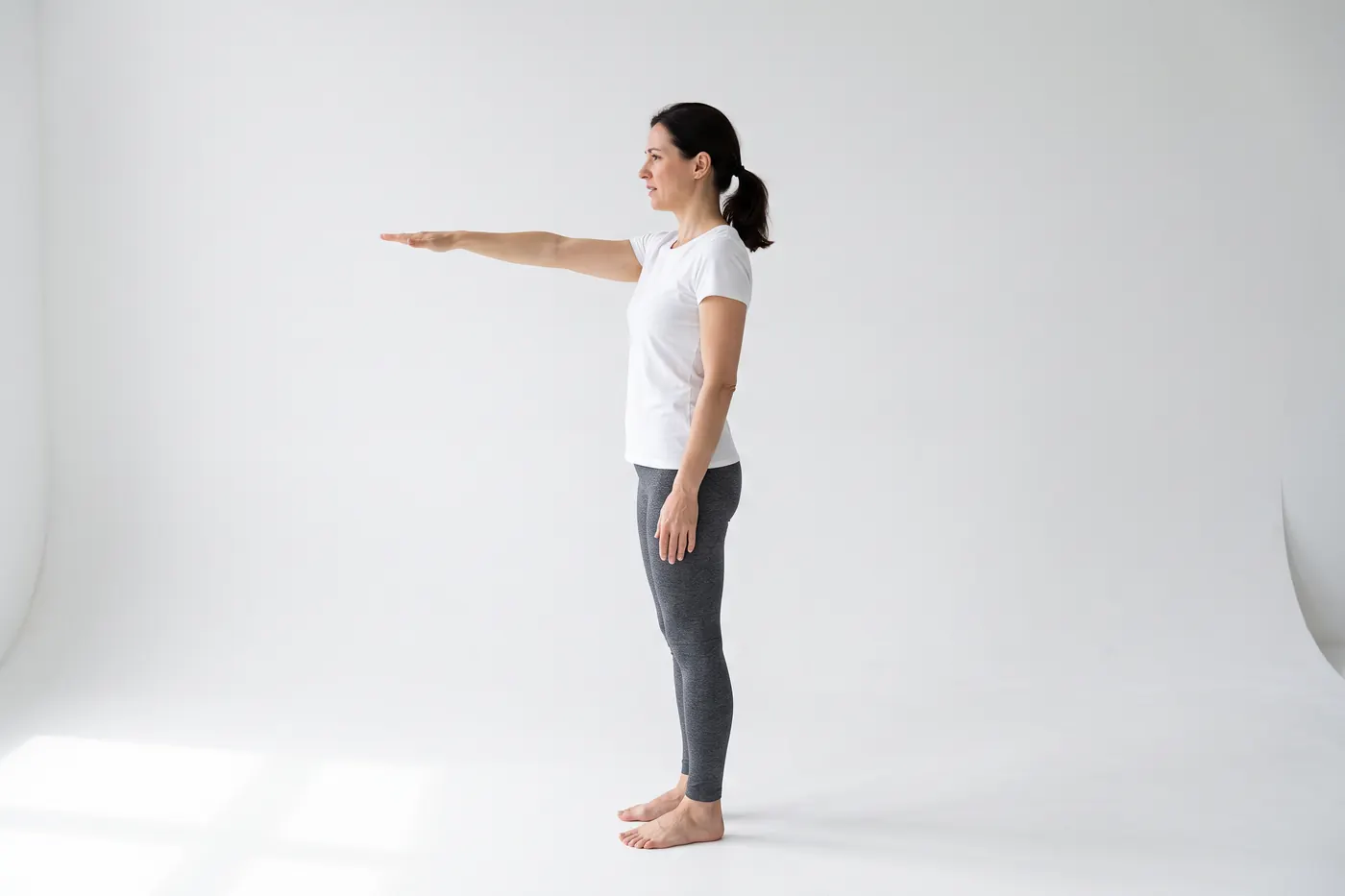 Side view of a woman in a white studio lifting one arm straight out in front of her to shoulder height, her other arm relaxed at her side
