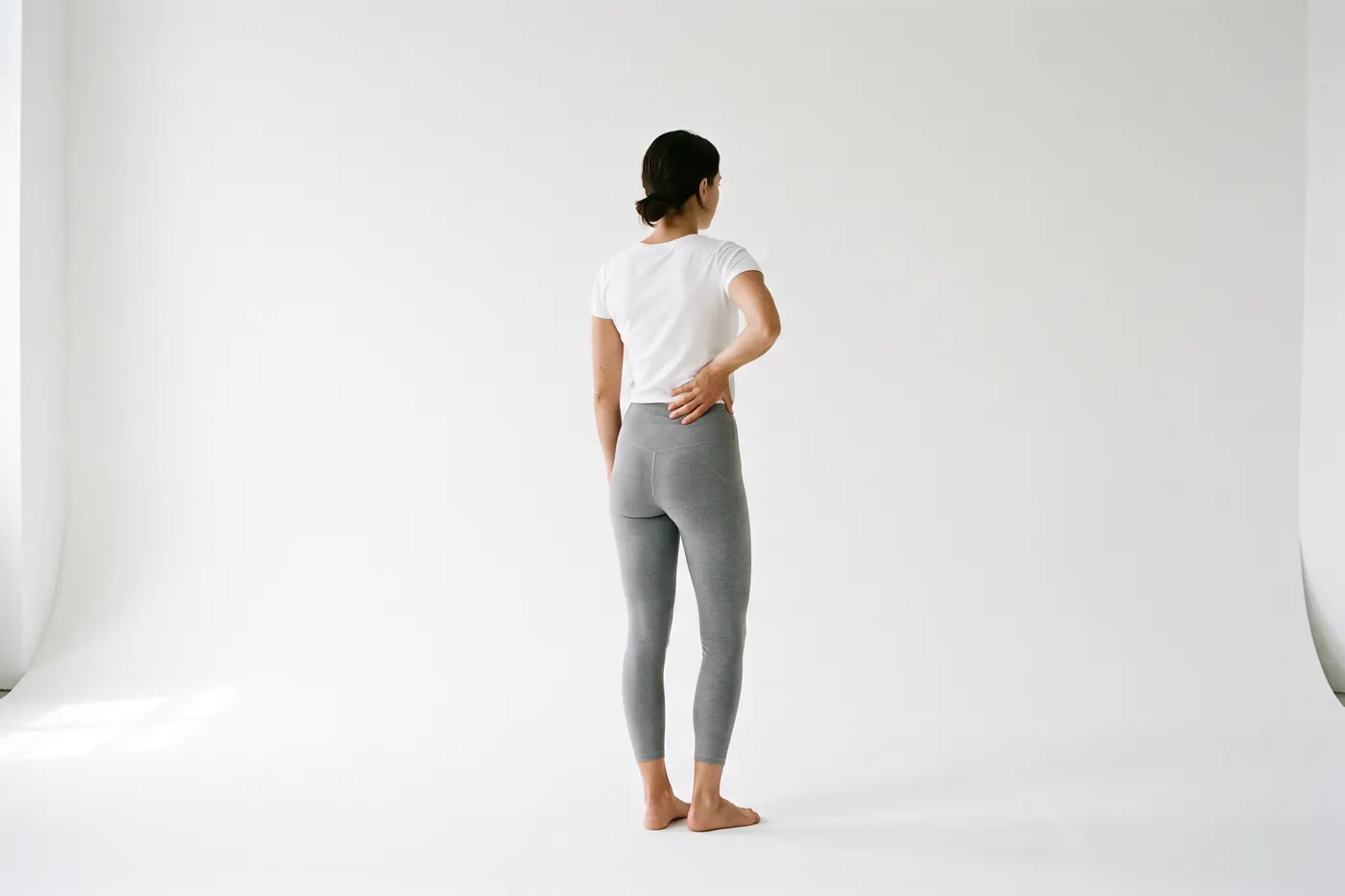 Back view of a woman in a white studio, one hand pressed to her lower back