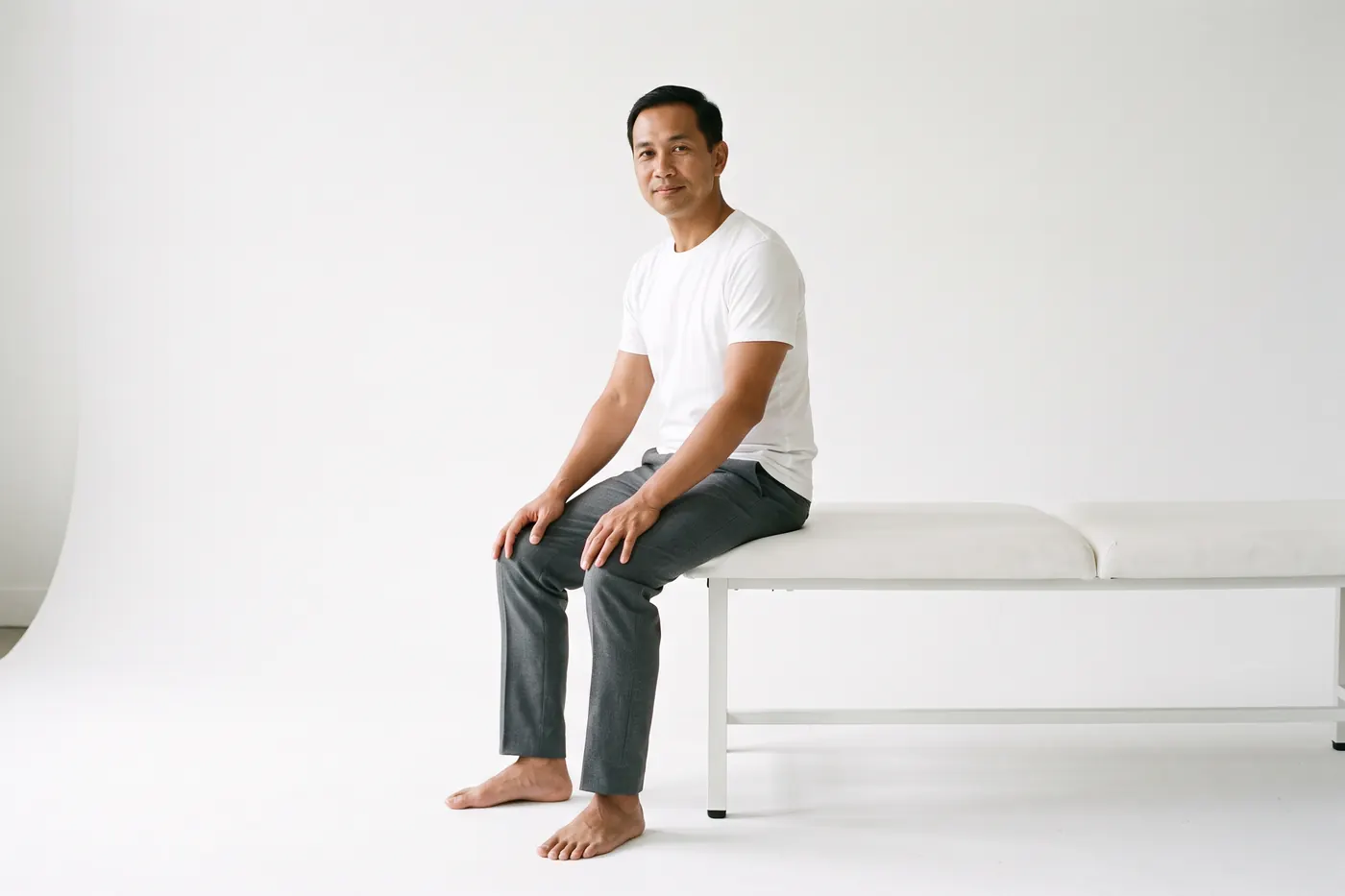 A calm man seated on the edge of a treatment table in a white studio, hands resting on his knees, turned three-quarters toward the camera