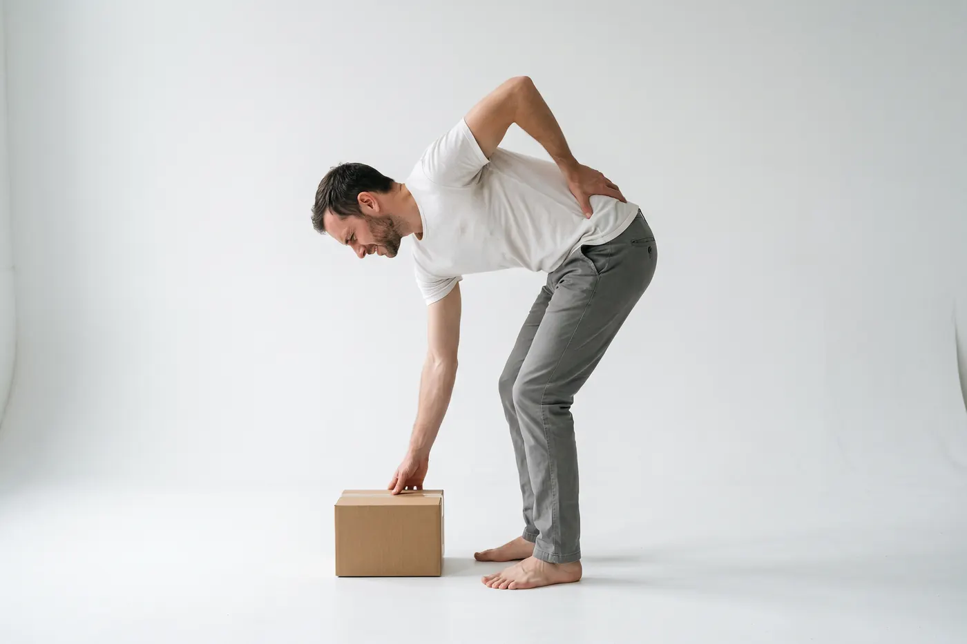 Side view of a man in a white studio bending to pick up a small cardboard box, pausing halfway with one hand pressed to his lower back