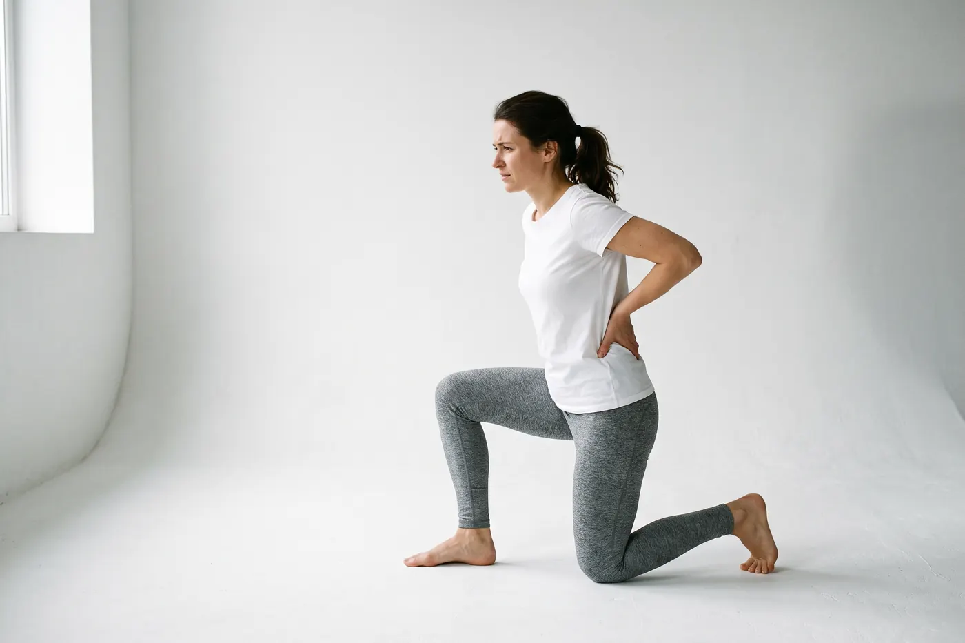 Side view of a woman in a white studio kneeling on one knee, one hand pressed to the side of her lower back