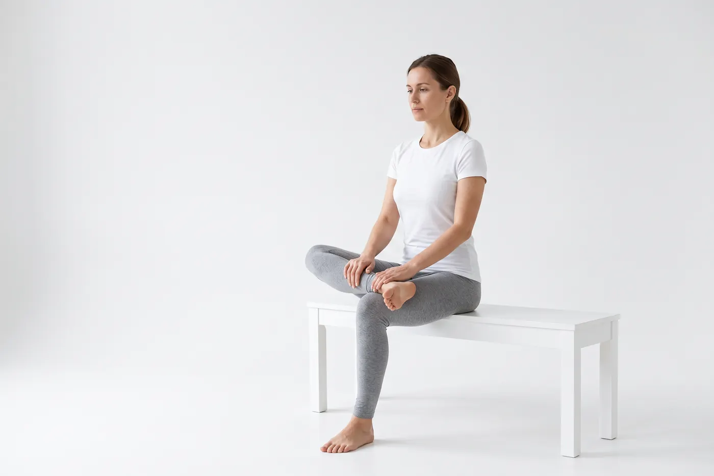 A woman seated in a white studio doing a figure-four stretch, one ankle resting across the opposite knee