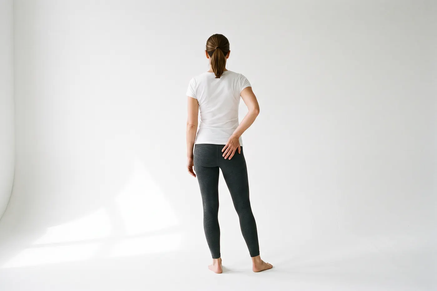 Back view of a woman in a white studio standing with her weight on one leg, one hand pressed low on the back of her pelvis