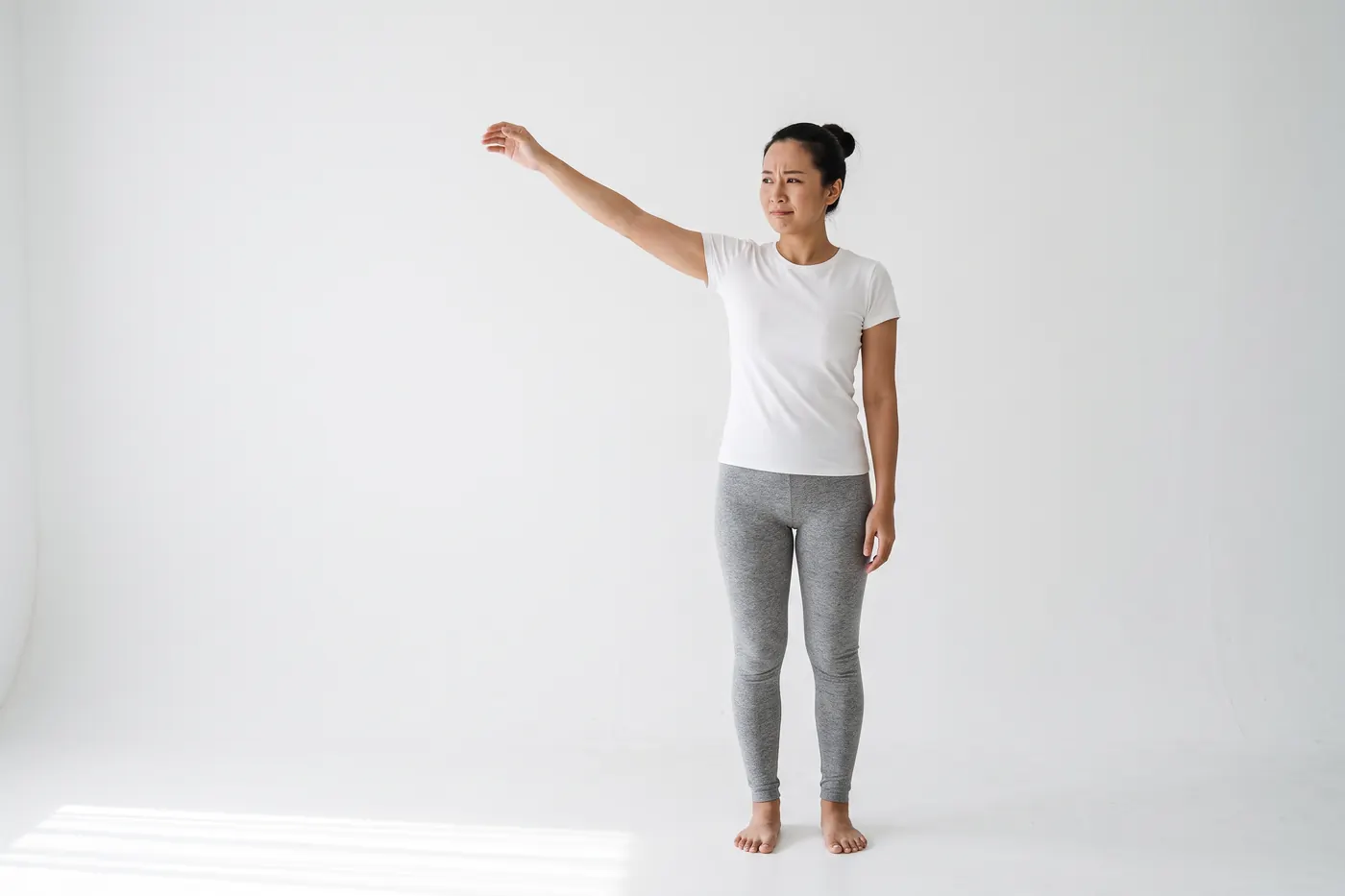 A woman in a white studio, seen from the front, raising one arm out to the side, her face showing mild discomfort partway through the lift