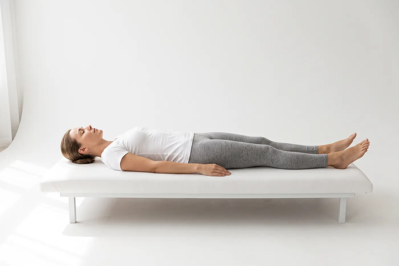 A woman lying face up on a padded treatment table in a white studio, relaxed, with her eyes closed