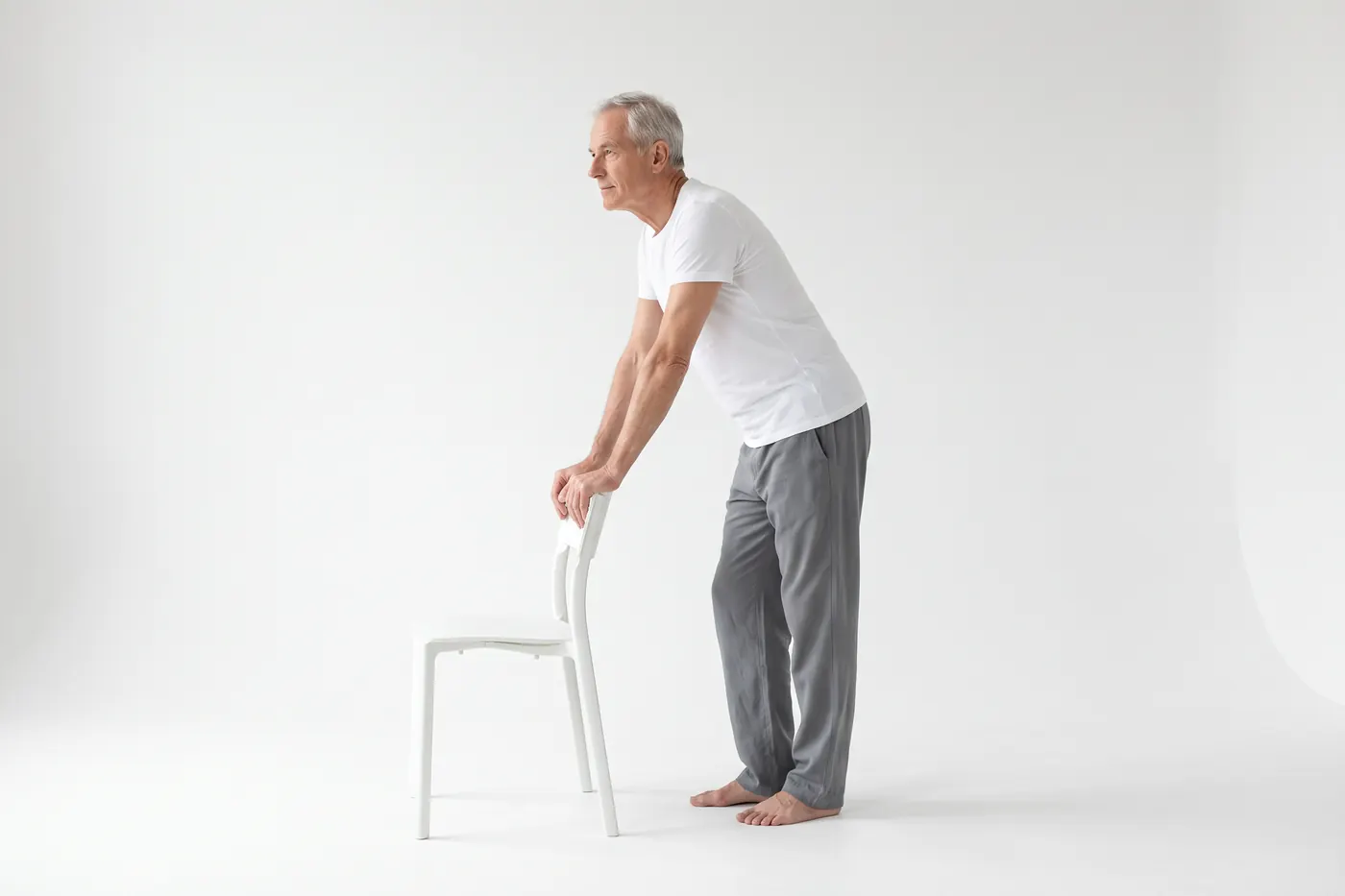 An older man in a white studio seen from the side, leaning forward slightly with both hands on the back of a chair