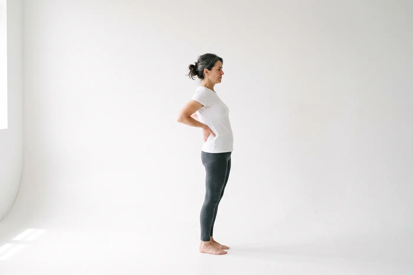 Side view of a woman in a white studio standing with both hands supporting the small of her back