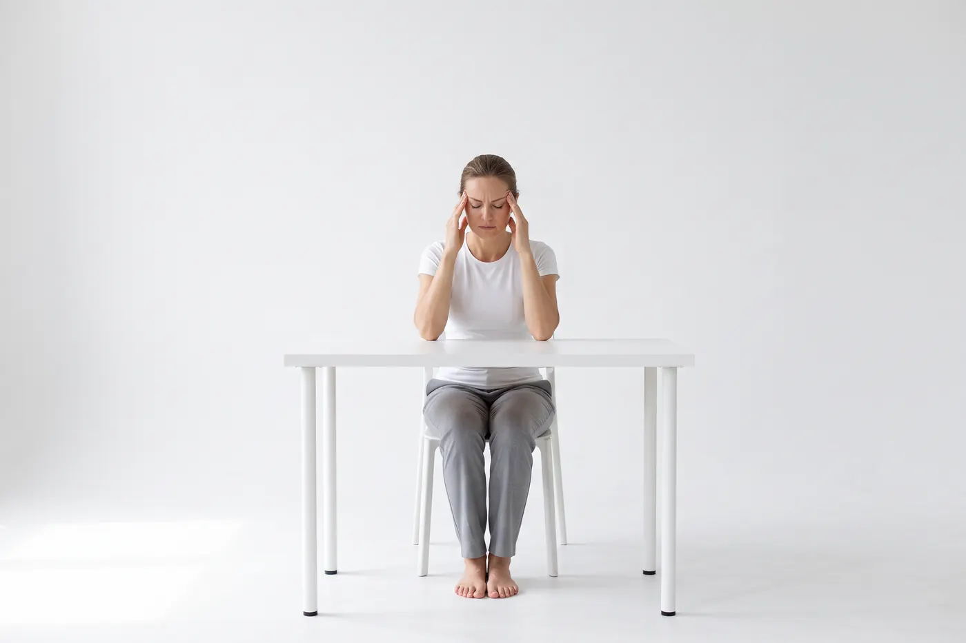 A woman seated at a desk in a white studio, eyes closed, fingertips pressed to her temples