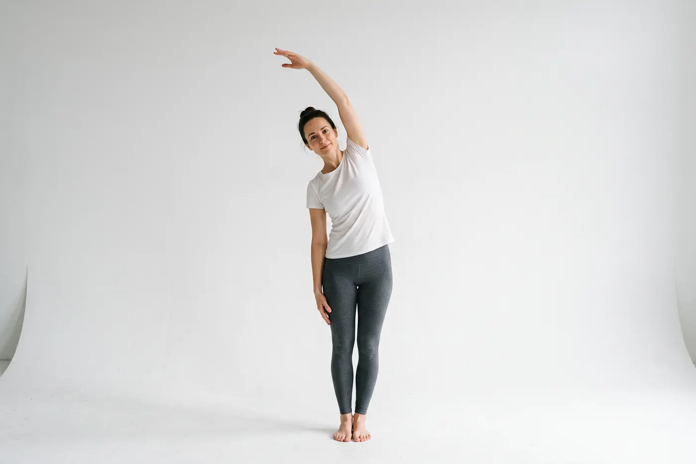 Front view of a woman in a white studio in a slow standing side stretch, one arm reaching up and over her head
