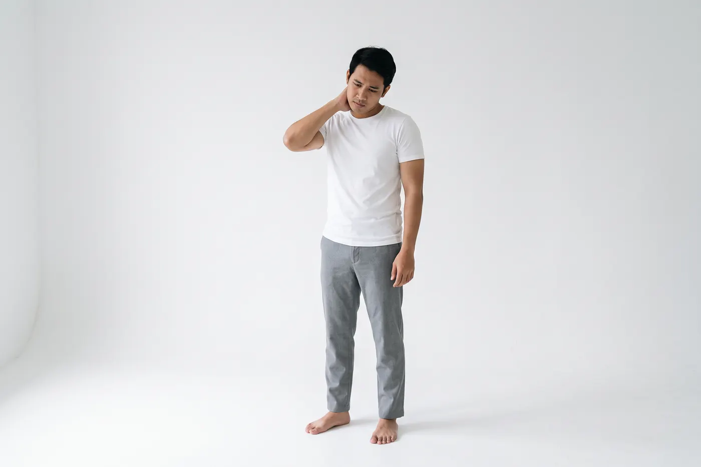 Front view of a man in a white studio holding the back of his neck with one hand, his head tilted slightly